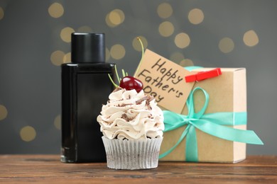 Photo of Greeting card with phrase Happy Father's Day, cupcake, gift box and perfume on wooden table against blurred lights