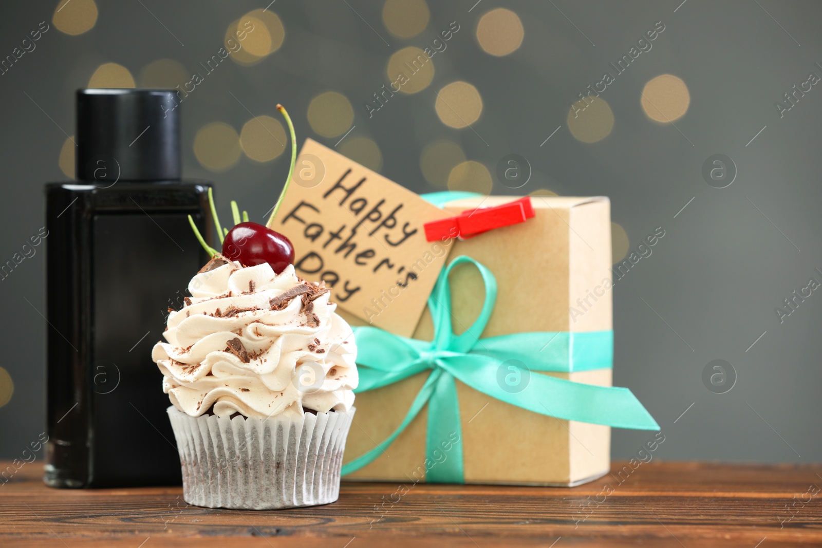 Photo of Greeting card with phrase Happy Father's Day, cupcake, gift box and perfume on wooden table against blurred lights