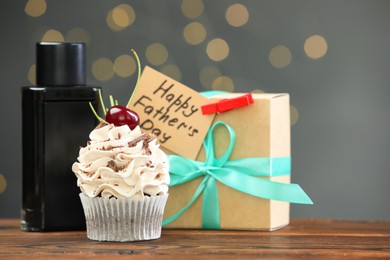 Greeting card with phrase Happy Father's Day, cupcake, gift box and perfume on wooden table against blurred lights