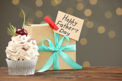 Photo of Greeting card with phrase Happy Father's Day, cupcake and gift box on wooden table against blurred lights. Space for text