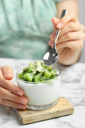 Woman eating tasty yogurt with fresh kiwi at white marble table, closeup