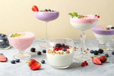 Photo of Different tasty yogurts with fresh berries and granola in glass dishware on gray textured table against pale yellow background