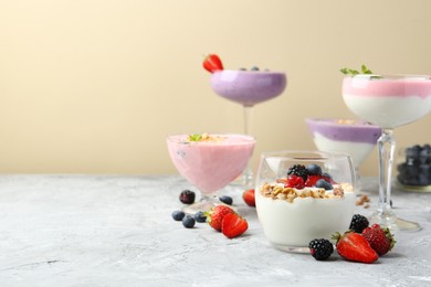 Photo of Different tasty yogurts with fresh berries and granola in glass dishware on gray textured table against pale yellow background, space for text