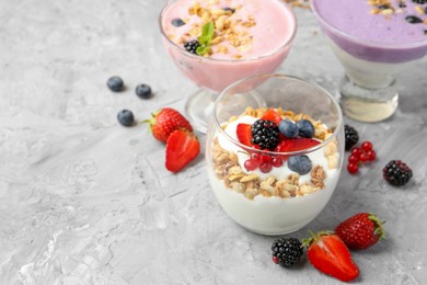 Photo of Different tasty yogurts with fresh berries and granola in glass dessert bowls on gray textured table, closeup. Space for text