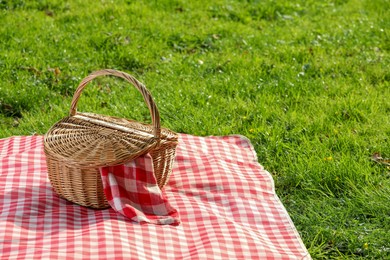 Photo of One picnic wicker basket with checkered napkin and blanket on green grass. Space for text