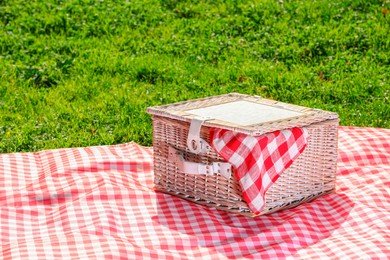 Photo of One picnic wicker basket with checkered napkin and blanket on green grass. Space for text