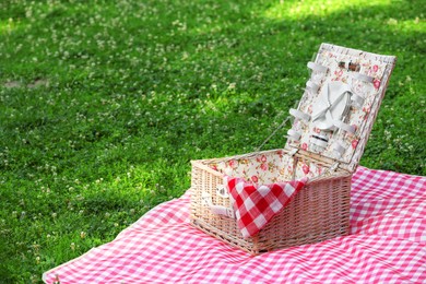 Photo of One picnic wicker basket with checkered napkin, tableware and blanket on green grass. Space for text