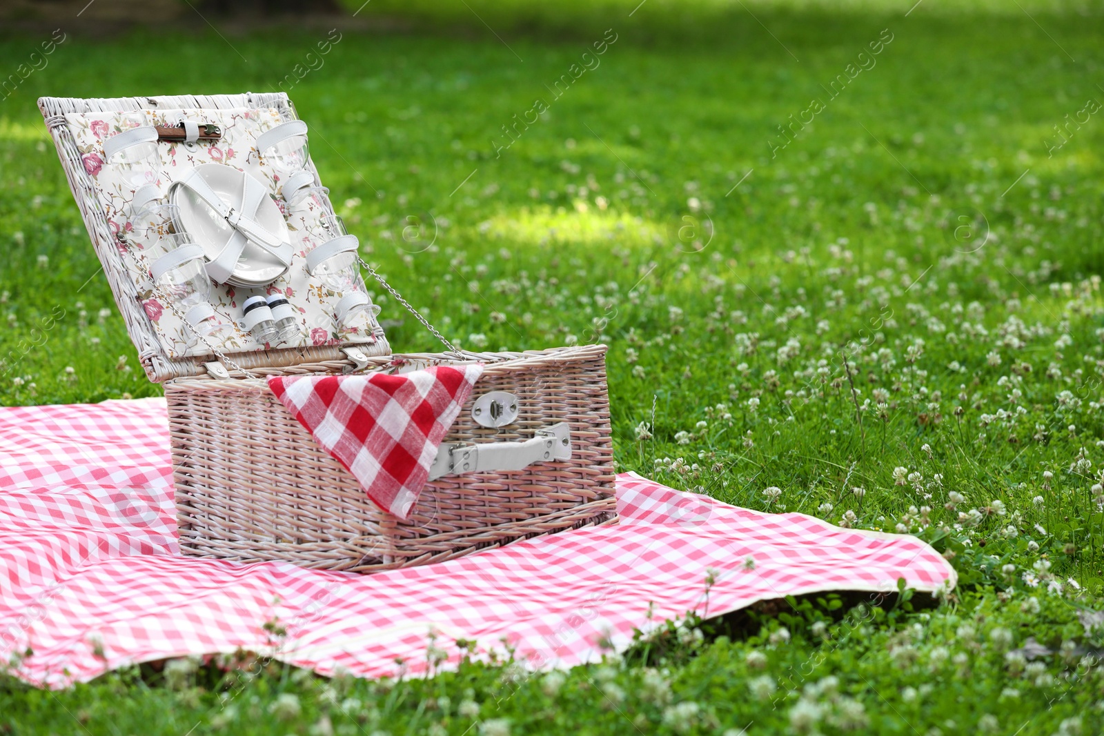 Photo of One picnic wicker basket with checkered napkin, tableware and blanket on green grass. Space for text