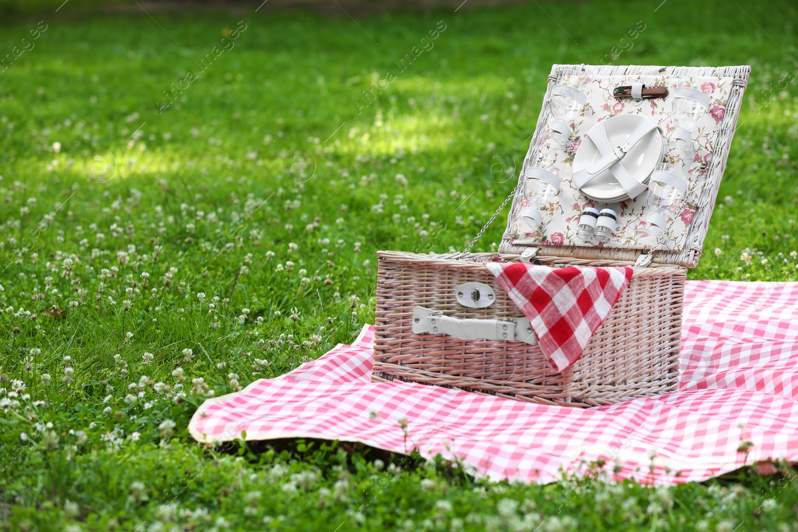 Photo of One picnic wicker basket with checkered napkin, tableware and blanket on green grass. Space for text