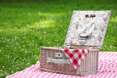 Photo of One picnic wicker basket with checkered napkin, tableware and blanket on green grass. Space for text