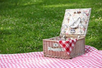 Photo of One picnic wicker basket with checkered napkin, tableware and blanket on green grass. Space for text