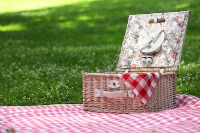 Photo of One picnic wicker basket with checkered napkin, tableware and blanket on green grass. Space for text