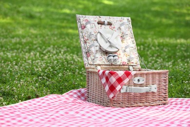 Photo of One picnic wicker basket with checkered napkin, tableware and blanket on green grass. Space for text