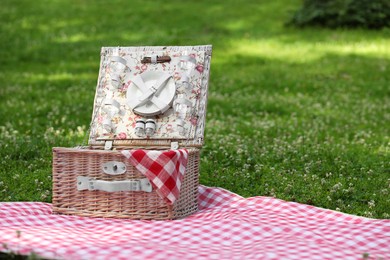 Photo of One picnic wicker basket with checkered napkin, tableware and blanket on green grass. Space for text