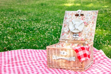 Photo of One picnic wicker basket with checkered napkin, tableware and blanket on green grass. Space for text