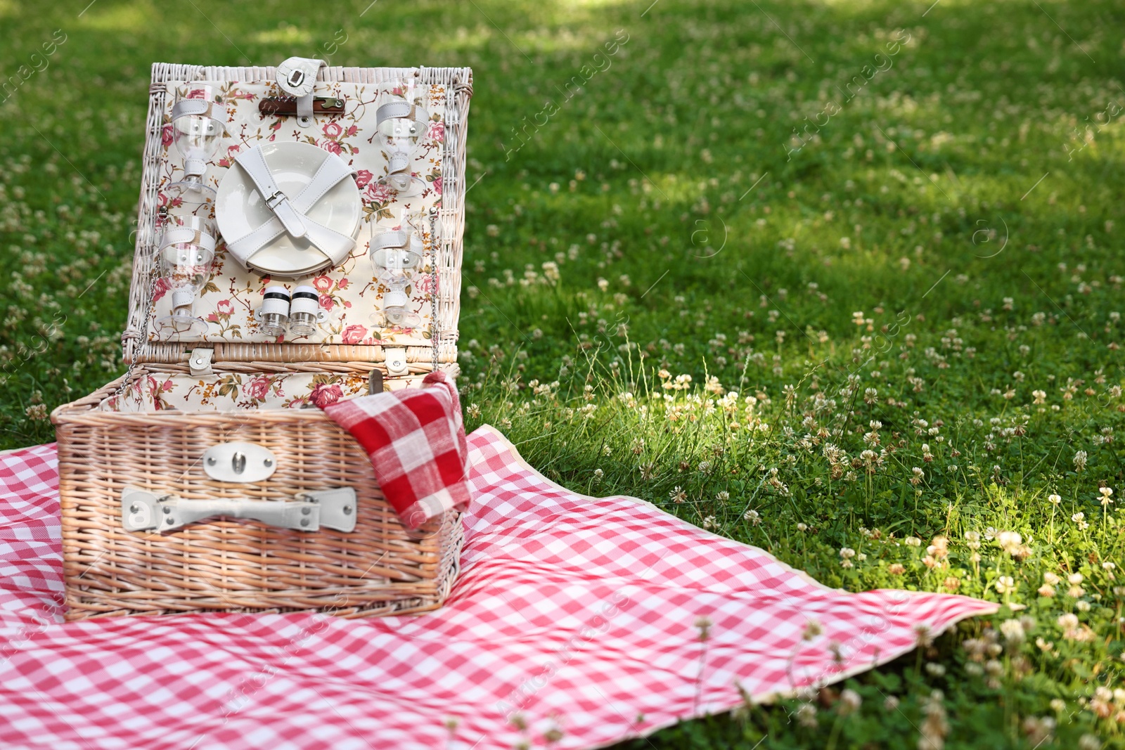 Photo of One picnic wicker basket with checkered napkin, tableware and blanket on green grass. Space for text