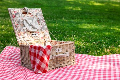 Photo of One picnic wicker basket with checkered napkin, tableware and blanket on green grass. Space for text