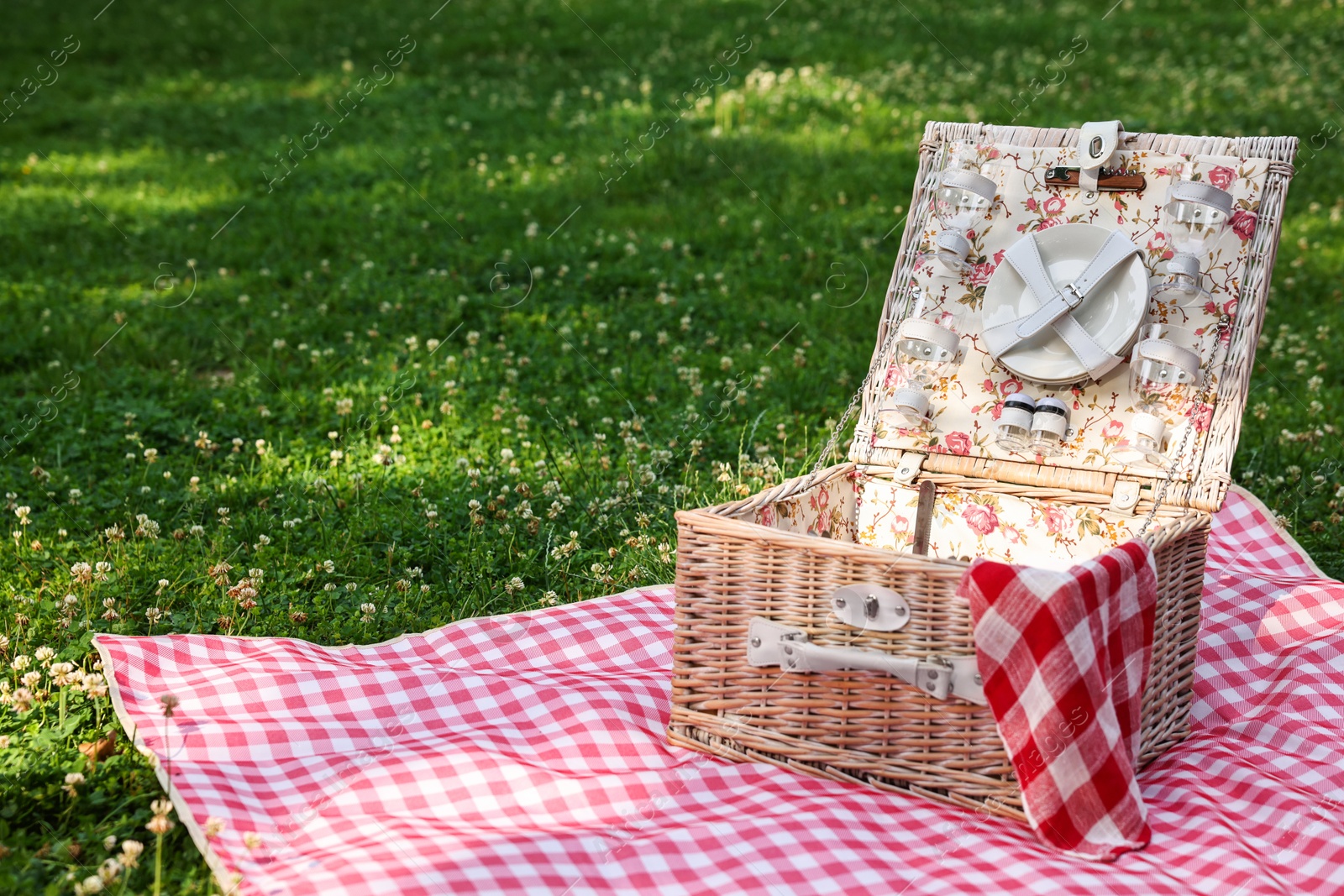 Photo of One picnic wicker basket with checkered napkin, tableware and blanket on green grass. Space for text