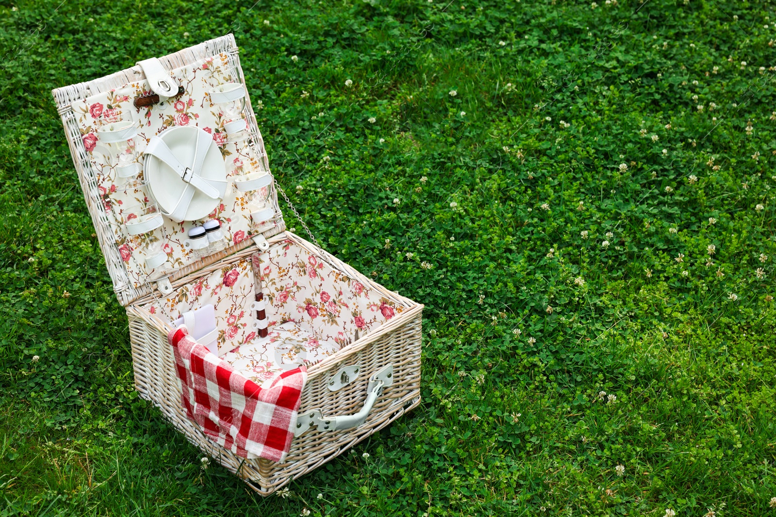 Photo of One picnic wicker basket with checkered napkin and tableware on green grass. Space for text