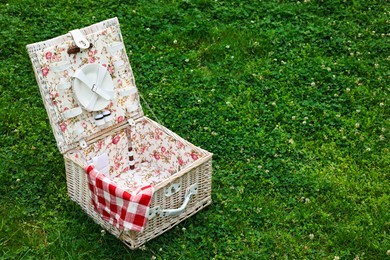 Photo of One picnic wicker basket with checkered napkin and tableware on green grass. Space for text