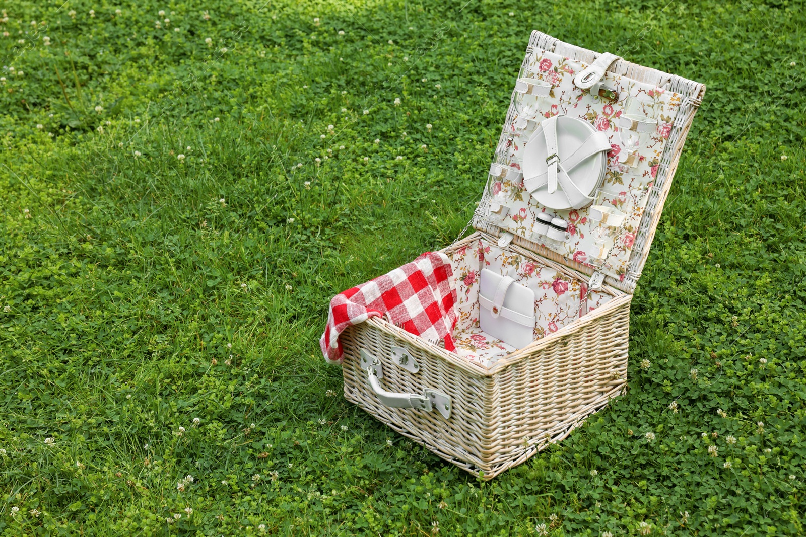 Photo of One picnic wicker basket with checkered napkin and tableware on green grass. Space for text