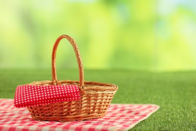 Photo of One picnic wicker basket with checkered napkin and blanket on grass against blurred green background, closeup. Space for text