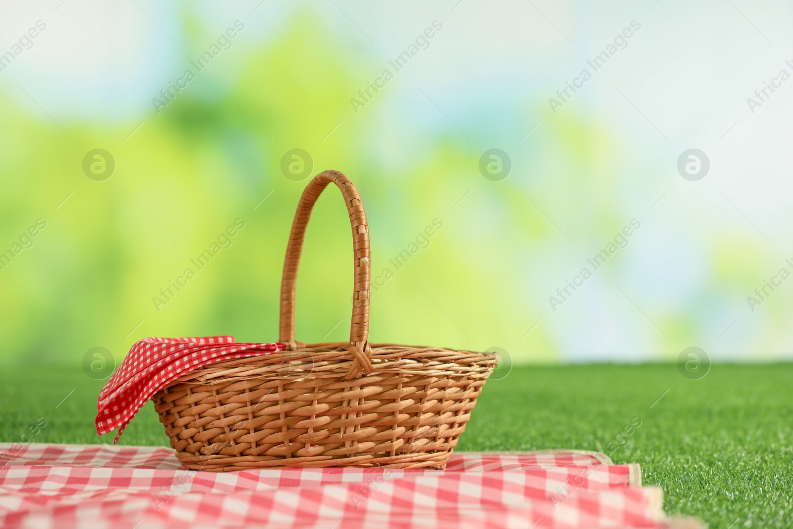 Photo of One picnic wicker basket with checkered napkin and blanket on grass against blurred green background. Space for text