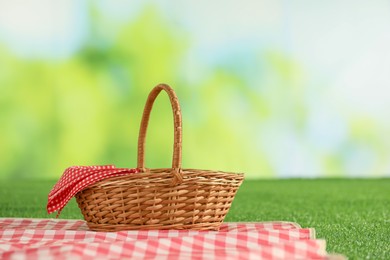 Photo of One picnic wicker basket with checkered napkin and blanket on grass against blurred green background. Space for text