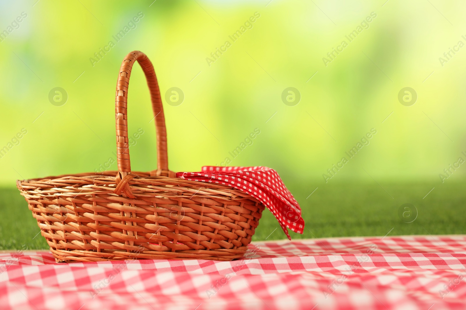 Photo of One picnic wicker basket with checkered napkin and blanket on grass against blurred green background. Space for text