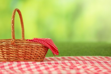 Photo of One picnic wicker basket with checkered napkin and blanket on grass against blurred green background. Space for text