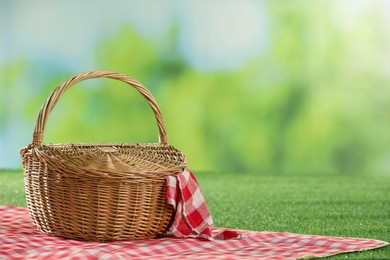 Photo of One picnic wicker basket with checkered napkin and blanket on grass against blurred green background. Space for text