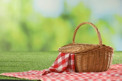 Photo of One picnic wicker basket with checkered napkin and blanket on grass against blurred green background. Space for text