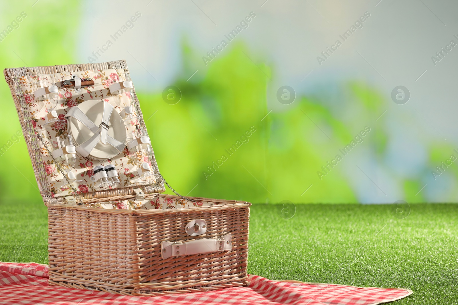 Photo of One picnic wicker basket with checkered napkin, tableware and blanket on grass against blurred green background. Space for text