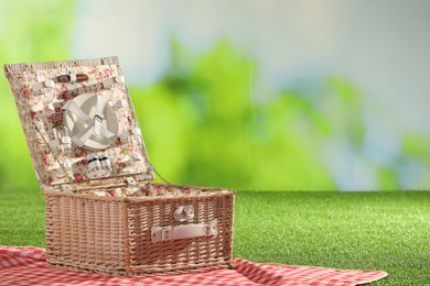 Photo of One picnic wicker basket with checkered napkin, tableware and blanket on grass against blurred green background. Space for text