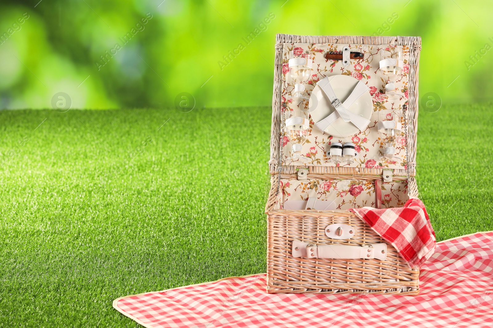 Photo of One picnic wicker basket with checkered napkin, tableware and blanket on grass against blurred green background. Space for text