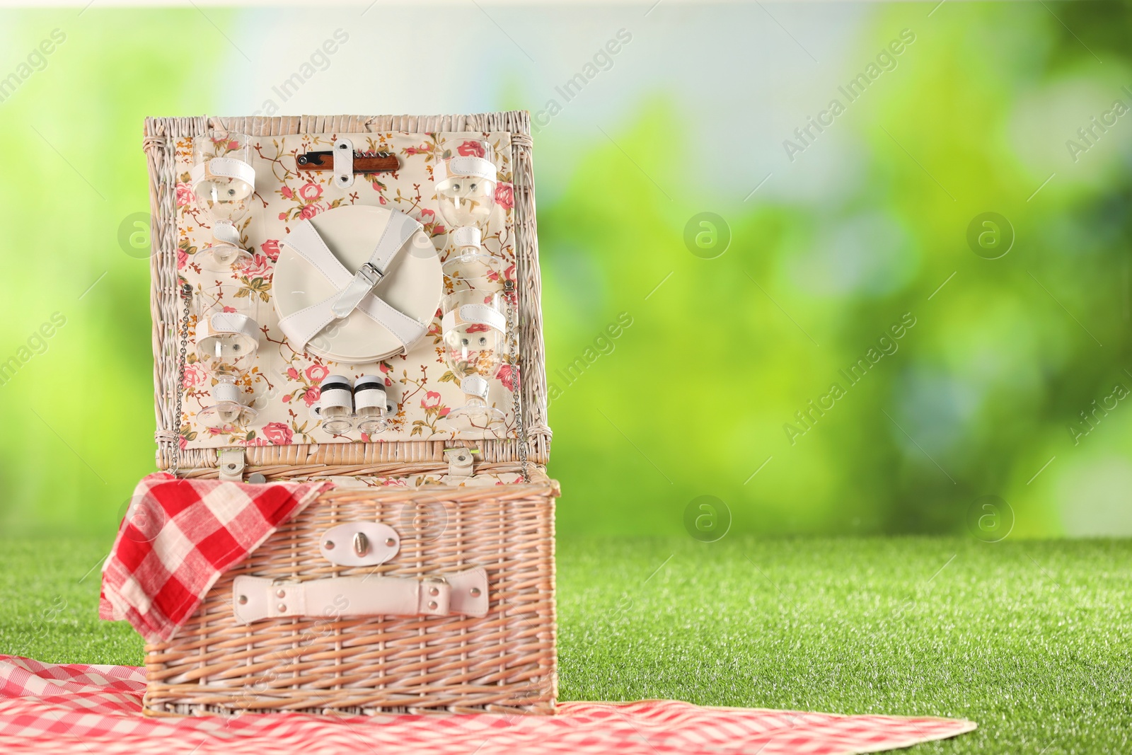 Photo of One picnic wicker basket with checkered napkin, tableware and blanket on grass against blurred green background. Space for text