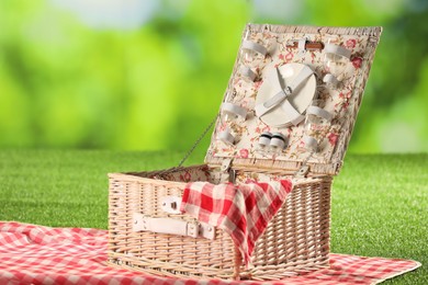 Photo of Picnic wicker basket with checkered napkin, tableware and blanket on grass against blurred green background, closeup. Space for text