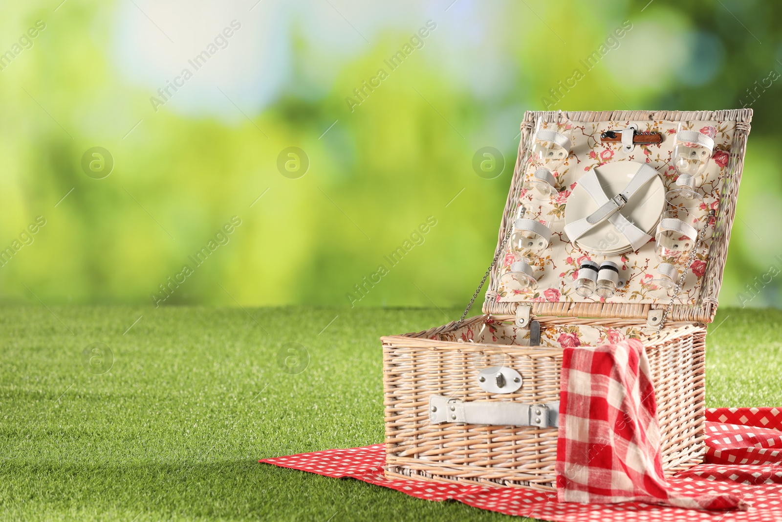 Photo of One picnic wicker basket with checkered napkin, tableware and blanket on grass against blurred green background. Space for text