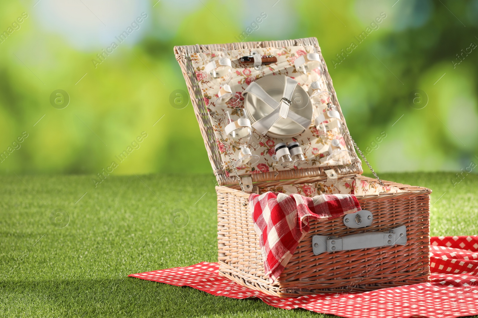 Photo of One picnic wicker basket with checkered napkin, tableware and blanket on grass against blurred green background. Space for text