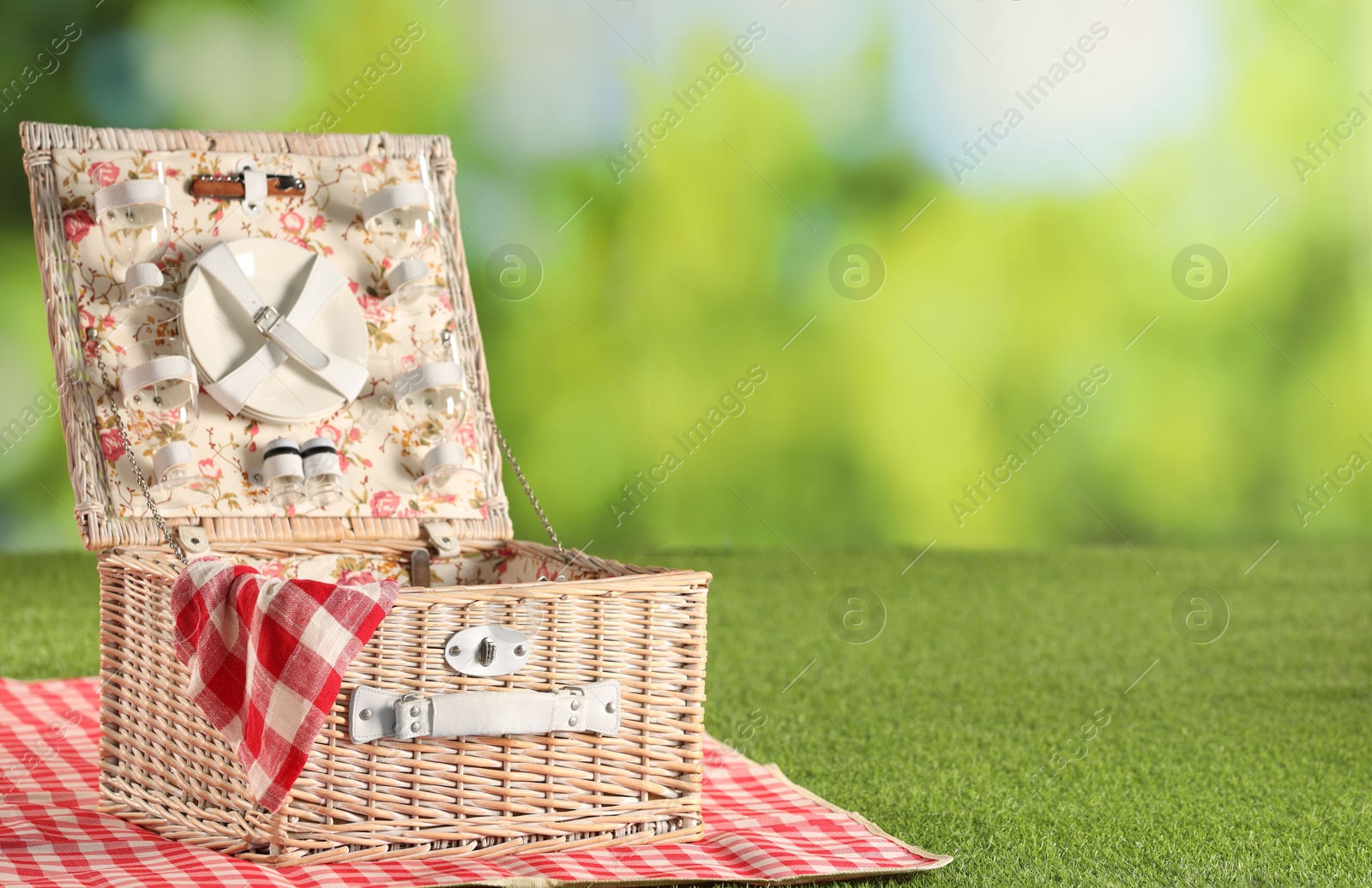 Photo of One picnic wicker basket with checkered napkin, tableware and blanket on grass against blurred green background. Space for text