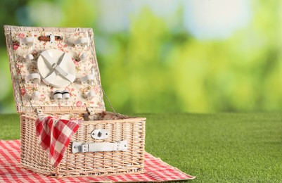 Photo of One picnic wicker basket with checkered napkin, tableware and blanket on grass against blurred green background. Space for text