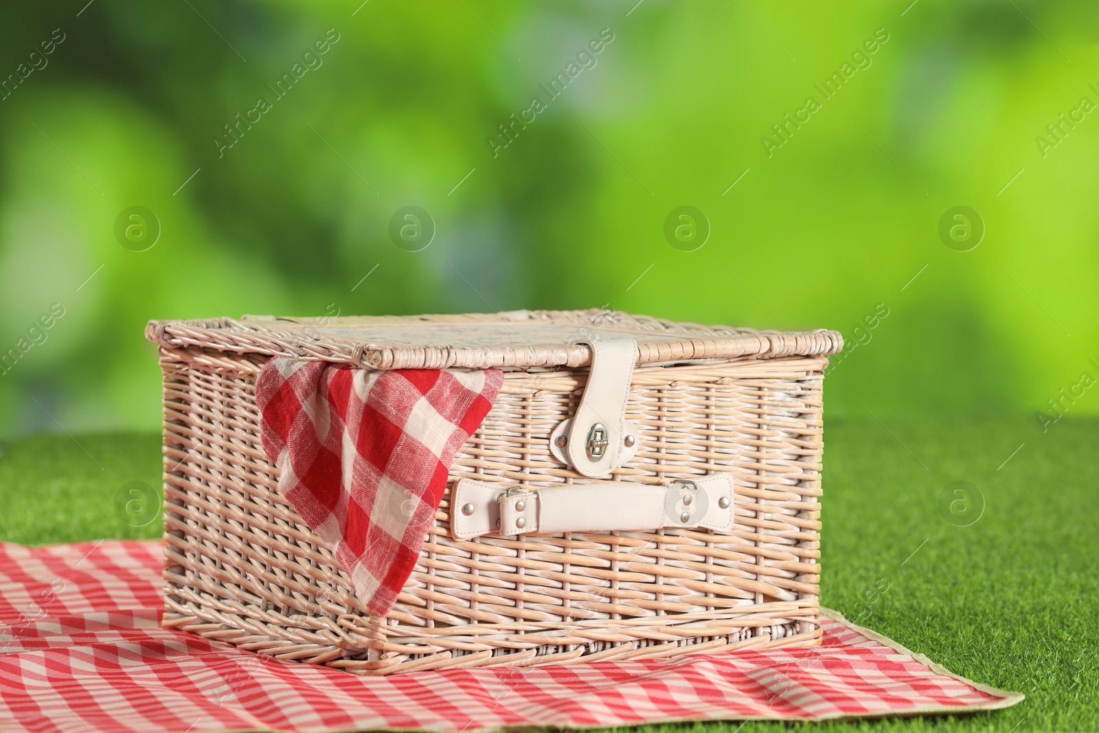 Photo of One picnic wicker basket with checkered napkin and blanket on grass against blurred green background, closeup