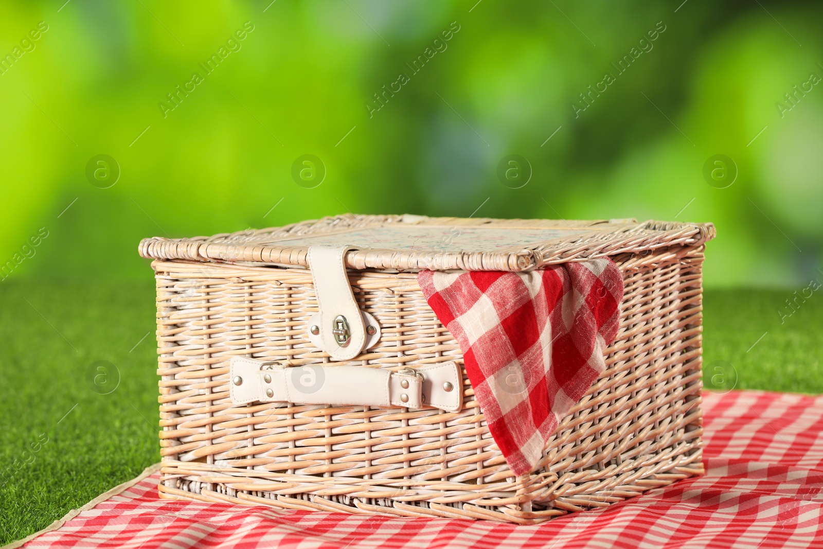 Photo of One picnic wicker basket with checkered napkin and blanket on grass against blurred green background, closeup