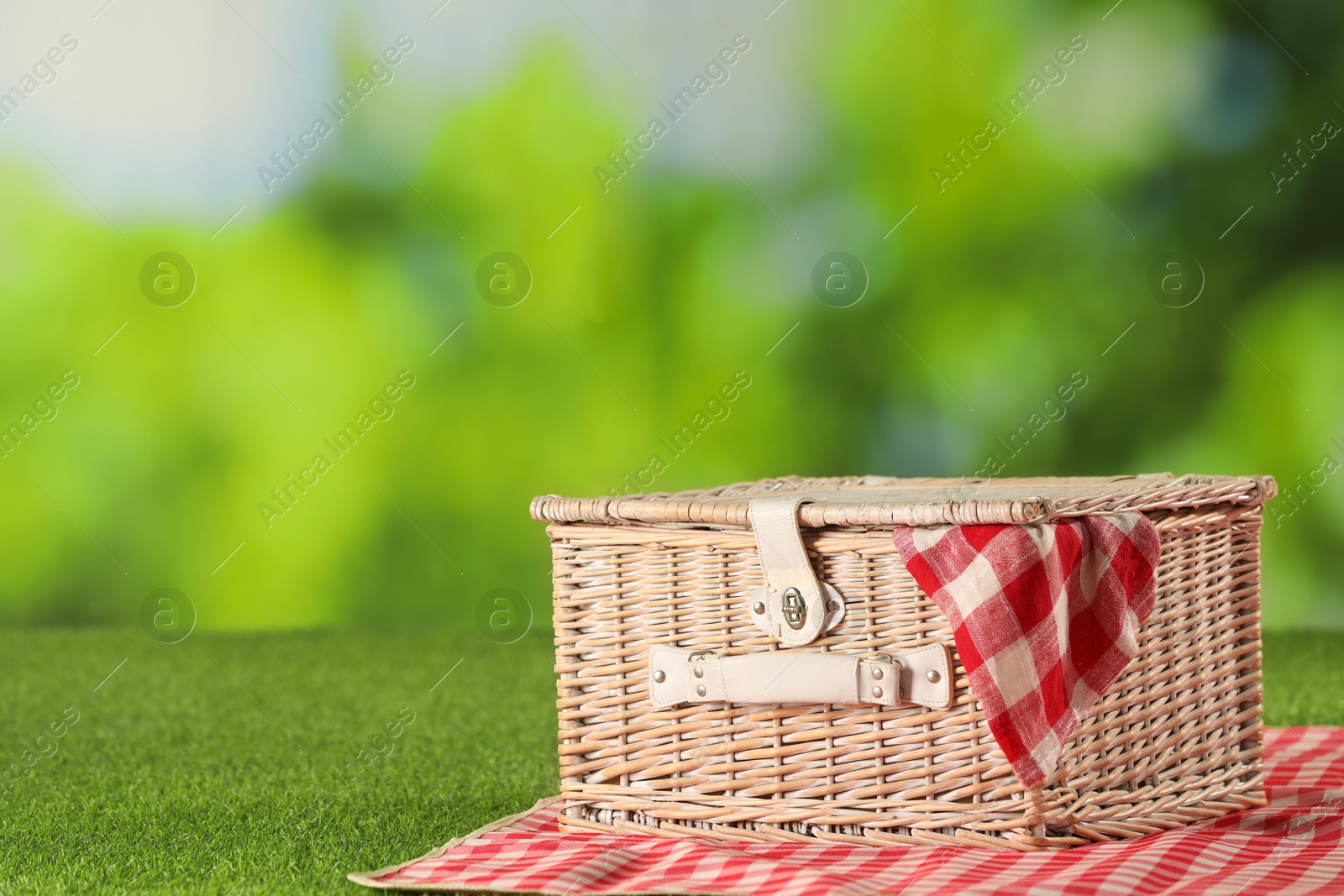 Photo of One picnic wicker basket with checkered napkin and blanket on grass against blurred green background, closeup. Space for text