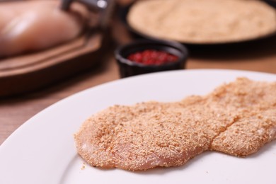 Making schnitzel. Plate with raw slice of meat in bread crumbs on wooden table, closeup