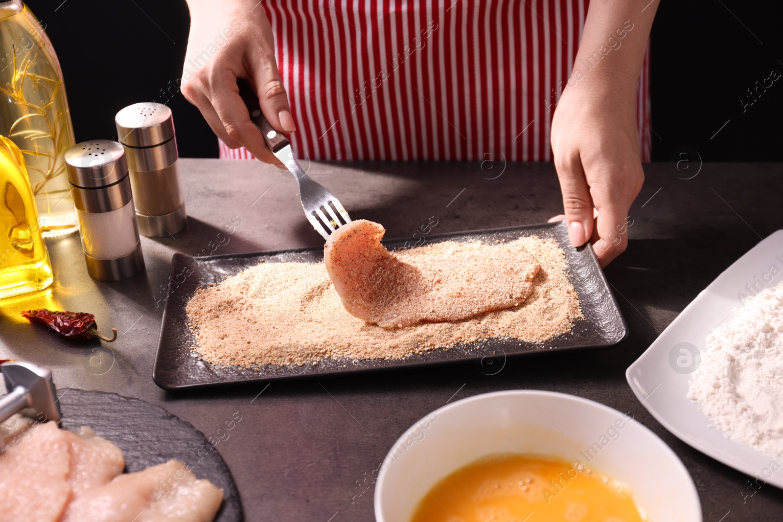 Photo of Making schnitzel. Woman coating slice of meat with bread crumbs at dark table, closeup