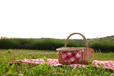 Photo of Picnic wicker basket with napkin and red checkered blanket on green grass outdoors, space for text