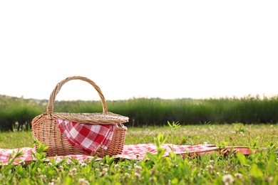 Photo of Picnic wicker basket with napkin and red checkered blanket on green grass outdoors, space for text