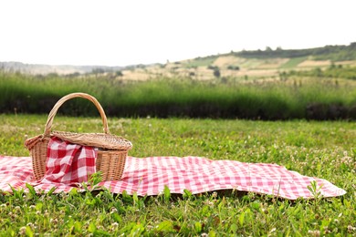 Photo of Picnic wicker basket with napkin and red checkered blanket on green grass outdoors, space for text