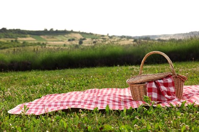 Photo of Picnic wicker basket with napkin and red checkered blanket on green grass outdoors, space for text